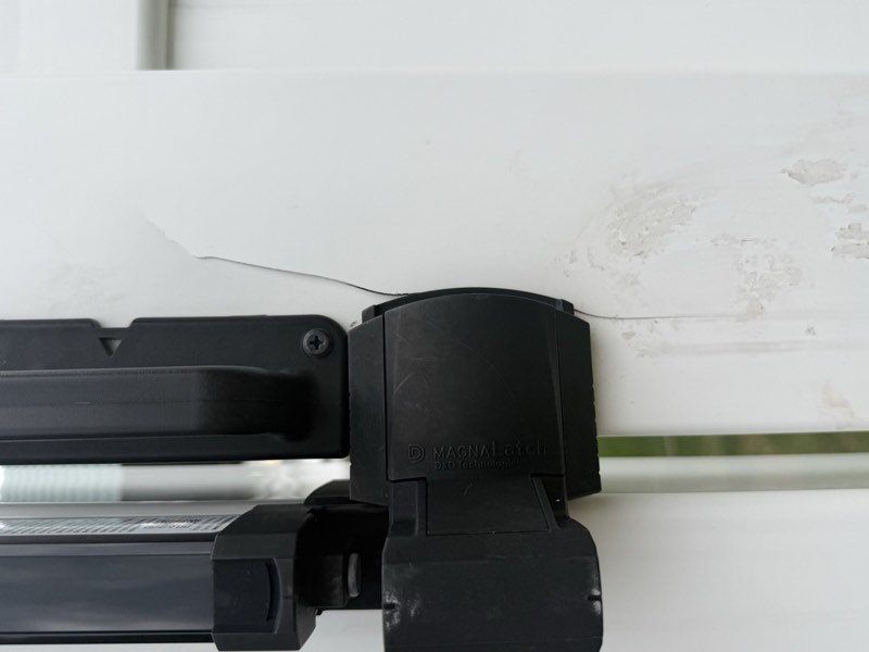 A crack running out of a latch fastener on a gate, beside a black MagnaLatch pool safety latch, with caulk smeared below.